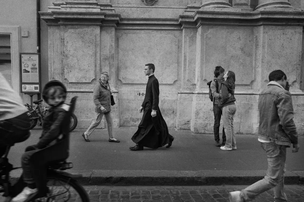 Pedestrians pass a stone facade while a scooter crosses the foreground
