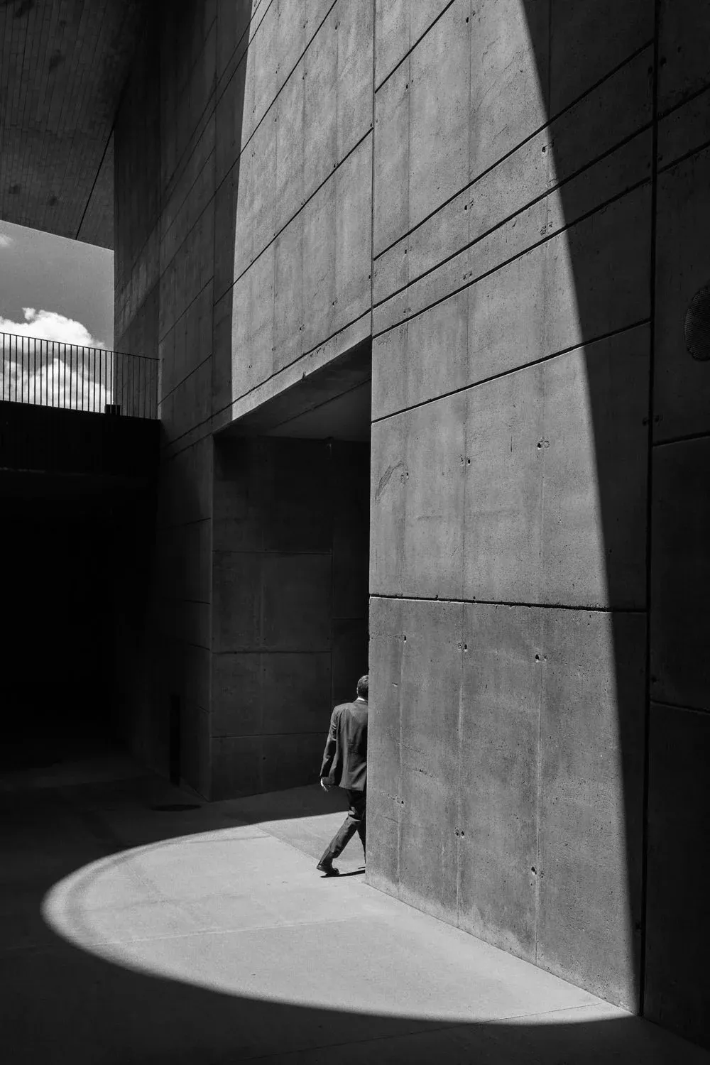 A pedestrian emerges beside concrete walls and a curved patch of sunlight